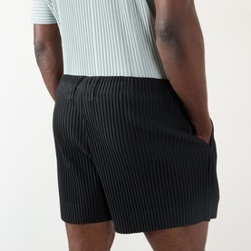 Model wearing Issey Miyake black pleated polyester shorts, styled with a mint ribbed T-shirt, hands in pockets, rear view.