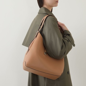 Model carrying Kate Spade Deco Large Shoulder Bag in Garden Soil, grained leather with gold-tone hardware, worn on shoulder over olive trench coat