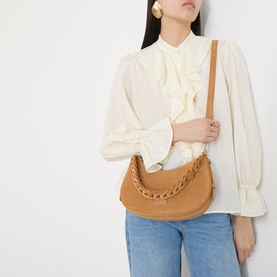 Model carrying Kate Spade Liv Suede Shoulder Bag in Garden Soil, with braided strap and gold-tone hardware, styled with white ruffle blouse and jeans