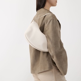 Model carrying Kate Spade Loop Pebbled Leather Shoulder Bag in Cashew Milk, gold-tone hardware, worn over shoulder with taupe suede jacket