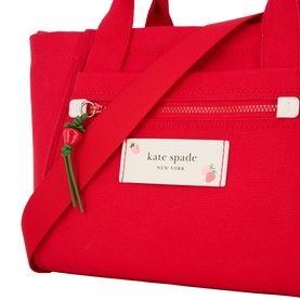 Kate Spade Dash Strawberry Canvas Tote Bag in Ruby Red, close-up of front zip pocket, logo badge, and strawberry charm detail