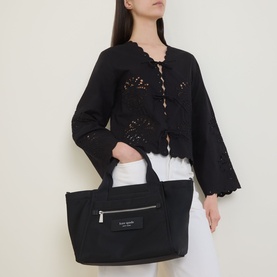 Model carrying Kate Spade Dash Nylon Tote Bag in black with silver-tone hardware and front zip pocket, styled with black lace top and white trousers.
