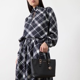 Model carrying MMK Quinn Small East West Tote Bag in black leather with gold-tone hardware and top handles, styled with black plaid dress.
