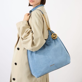 Model carrying MMK Cinch Large Messenger Bag in blue suede with gold-tone chain strap and branded key chain, styled with a beige trench coat