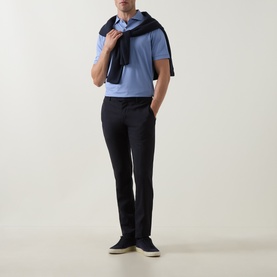 Model wearing Peter Millar Empire Performance Polo Shirt in Windy Blue with navy knitwear draped over shoulders, styled with black trousers and navy