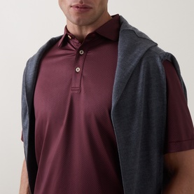 Model wearing Peter Millar bordeaux polo shirt with subtle texture, three-button placket, styled with a grey knitwear draped over shoulders.