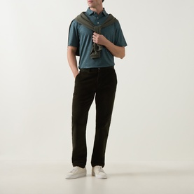 Model wearing Peter Millar polo in Pinebrook, styled with black trousers, white trainers, and an olive crossbody bag.