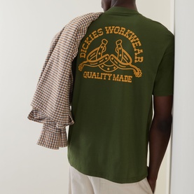 Model wearing Dickies Crew Neck T in Cypress green with orange logo graphic on back, styled with a checked shirt draped over shoulder.