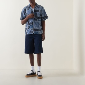 Model wearing Dickies rinsed navy cotton shorts, styled with a blue patchwork print short-sleeve shirt and low-top trainers.