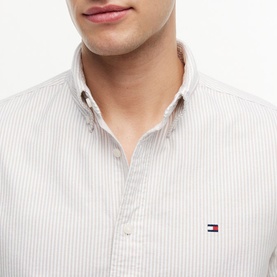 Model wearing Tommy Hilfiger shirt in Coastal Taupe stripe, close-up showing button-down collar and logo badge detail.