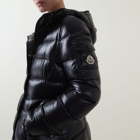 Model wearing Moncler black quilted hooded puffer jacket in high-shine polyamide with logo arm badge, hands in pockets.