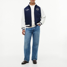 Model wearing Tommy Hilfiger Carbon Navy varsity bomber jacket with white sleeves and logo badge, styled with light-wash jeans