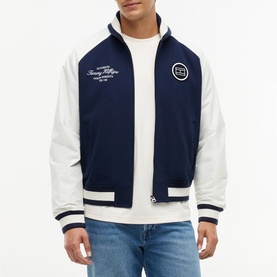 Model wearing Tommy Hilfiger Carbon Navy varsity bomber jacket with white sleeves, ribbed cuffs, and chest badge detail, styled with white crew-neck