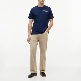 Model wearing Tommy Hilfiger Carbon Navy crew neck T-shirt with small chest logo, styled with beige straight-leg trousers and dark loafers.