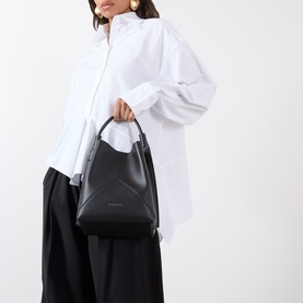 Model carrying Victoria Beckham Victoria Bucket Bag in black leather, top handle, gold-tone logo detail, styled with white shirt and black wide-leg