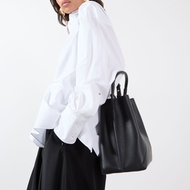 Model carrying Victoria Beckham Victoria Bucket Bag in black leather with top handle and gold-tone detailing, styled with white oversized shirt and