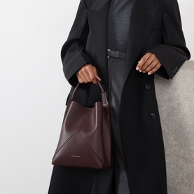 Model carrying Victoria Beckham Victoria Bucket Bag in dark burgundy leather, gold-tone logo, top handle, styled with black oversized coat