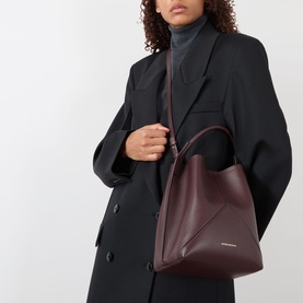 Model carrying Victoria Beckham Victoria Bucket Bag in dark burgundy leather with gold-foil logo, top handle and detachable shoulder strap, styled