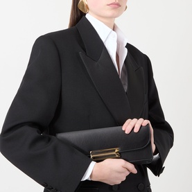 Model carrying Victoria Beckham Dorian East West Clutch in black satin with gold-tone hardware, styled with a black blazer and white shirt.