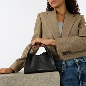 Model carrying Victoria Beckham Pli&eacute; Mini Crossbody Bag in black smooth Nappa leather with gold-tone hardware, held by top handle. Styled with blazer