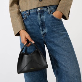Model carrying Victoria Beckham Pli&eacute; Mini Crossbody Bag in black smooth Nappa leather with gold-tone hardware, styled with blue jeans and tan blazer.