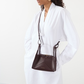 Model carrying Victoria Beckham Pli&eacute; Mini Crossbody Bag in dark burgundy smooth Nappa leather, gold-tone hardware, worn crossbody over white outfit
