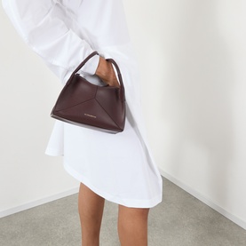 Model carrying Victoria Beckham Pli&eacute; Mini Crossbody Bag in dark burgundy smooth Nappa leather with gold-tone hardware and top handle, styled with