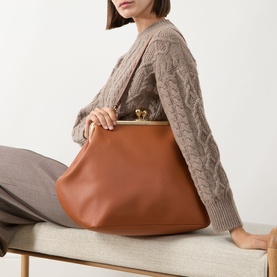 Model carrying Coach Kisslock Large Frame Bag in Amber Brown leather, gold-tone kisslock closure, styled with a chunky knit cardigan