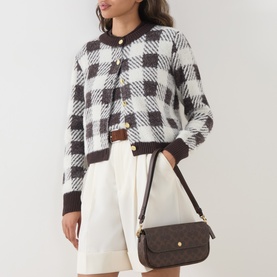 Model carrying Coach Brooke Chain Crossbody Bag in Maple, gold-tone hardware, C-logo print coated canvas, worn with checked cardigan and white shorts