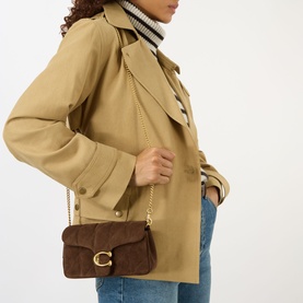 Model carrying Coach Tabby 19 Quilted Suede Crossbody Bag in Dark Chocolate with gold chain strap, styled with a beige jacket and jeans