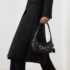 Model carrying Coach Ergo 26 Belted Shoulder Bag in black leather with gold-tone hardware and belted clasp, styled with a black coat and trousers
