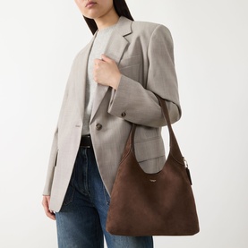 Model carrying Coach Brooklyn 28 Suede Shoulder Bag in Dark Chocolate with gold-toned hardware, styled with grey blazer and jeans