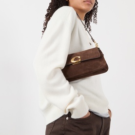 Model carrying Coach Jet Suede Shoulder Bag in dark chocolate with gold-toned C hardware and chain strap, styled with white knit top