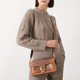 Model carrying Coach Tabby 26 Shoulder Bag in dark brown leather with gold-tone C-logo hardware, worn crossbody, styled with a cable-knit jumper