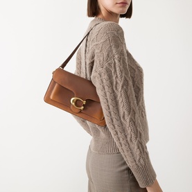 Model carrying Coach Tabby 26 Shoulder Bag in dark brown leather with gold-tone C-logo hardware, worn on shoulder, styled with beige cable-knit jumper