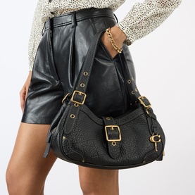Model carrying Coach Drifter Small Shoulder Bag in black pebbled leather with gold-tone hardware, buckle straps and padlock charm, styled with black