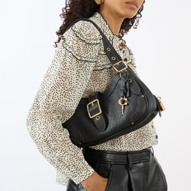 Model carrying Coach Drifter Small Shoulder Bag in black pebbled leather with gold-tone hardware, buckle detail and padlock charm, styled with a