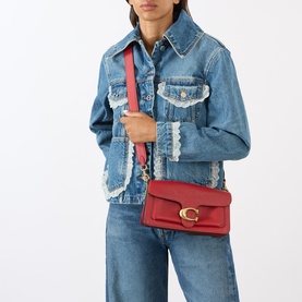 Model carrying Coach Tabby 26 Shoulder Bag in Poppy red leather with gold-tone C-logo hardware, worn crossbody over denim jacket and jeans