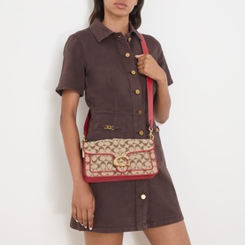 Model carrying Coach Tabby 26 Jacquard Shoulder Bag in Poppy Signature, gold-tone hardware, red contrast trim, worn crossbody over dark brown dress