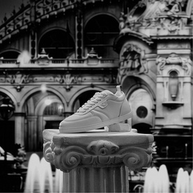 Christian Louboutin white low-top leather sneaker displayed on a stone column plinth against a grand architectural backdrop.