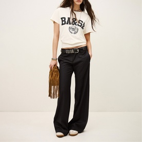 Model wearing Bash Womens French Dress in blue wide-leg trousers with cream logo tee and brown belt, carrying fringed suede bag