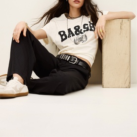 Model wearing Bash Womens French Dress in blue, styled with a black belt, black trousers, and white trainers, seated beside a wooden block.