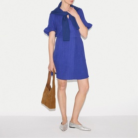 Model wearing Jigsaw blue short-sleeve mini dress with tie neck, styled with a tan tote bag and silver flat shoes.