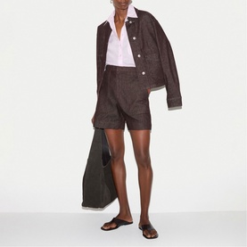 Model wearing dark brown linen shorts and matching jacket, styled with white shirt, brown slide sandals, carrying an olive tote bag