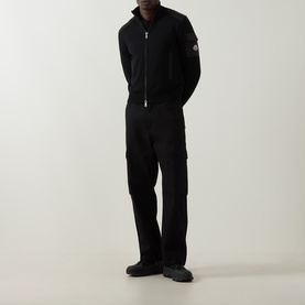 Model wearing Moncler black zip-up cardigan with logo badge, styled with matching black cargo trousers and dark chunky-sole shoes.