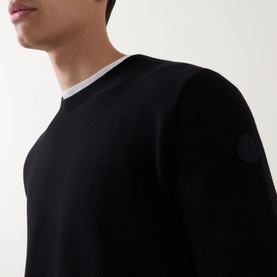 Model wearing black textured crew neck knit with white undershirt visible at collar and small logo badge on sleeve