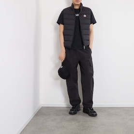 Model wearing Moncler Contrin Down Gilet in black, styled with black cargo trousers and a black T-shirt, holding a cap.