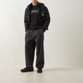 Model wearing Moncler black quilted down jacket with hood, styled with dark grey wide-leg trousers and black trainers.