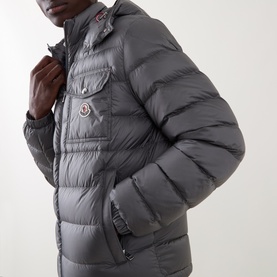 Model wearing Moncler grey quilted down puffer jacket with chest pocket, hood, and signature logo badge