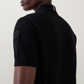 Model wearing Moncler black cotton polo shirt, close-up of sleeve badge logo and red-striped collar detail.